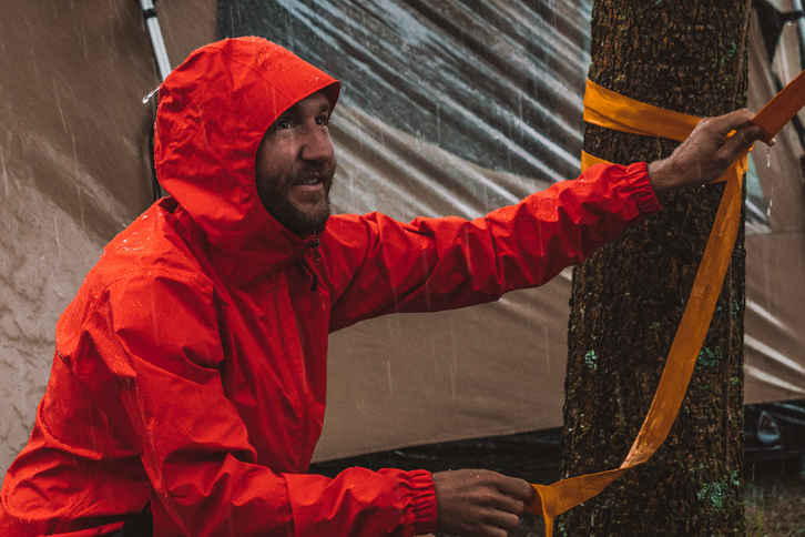 Person wearing a red rain jacket in the rain tying a tent to a tree.