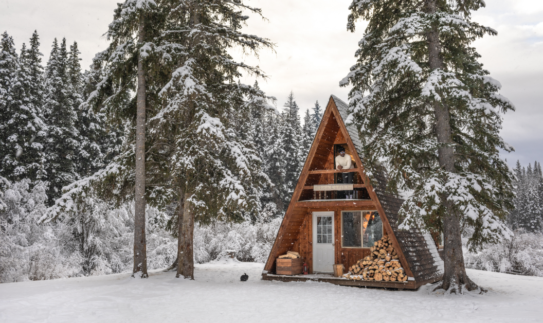 A small wooden cabin with a man inside surrounded by a cloudy recently snowed forest.