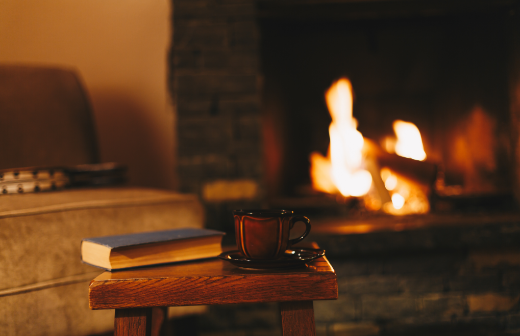 A book and cup of tea on a wooden stool with a fire lit in a dark brick fireplace in the background.