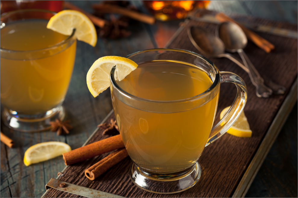A glass of hot toddy surrounded by slices of lemon and fresh cinnamon sticks.