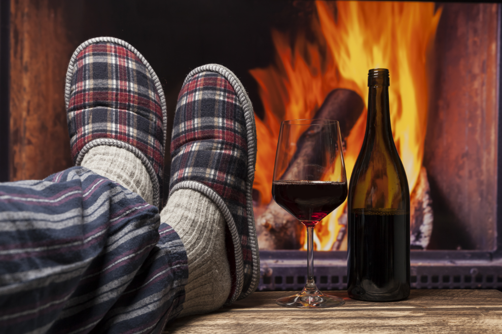 A person laying their feet in red plaid slippers in front of a lit fireplace next to a bottle and glass of red wine.