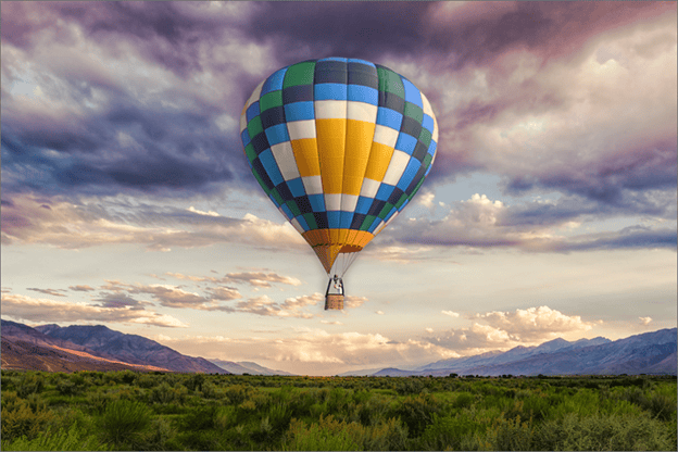 A multi-colored hot air balloon flying over a lush forest in the mountains during sunset.